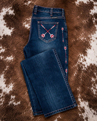 A pair of dark blue denim jeans laid flat on a brown and white cowhide rug. The back pocket features embroidered red and white floral and arrow details, and matching embroidery runs down the side seam of the leg.