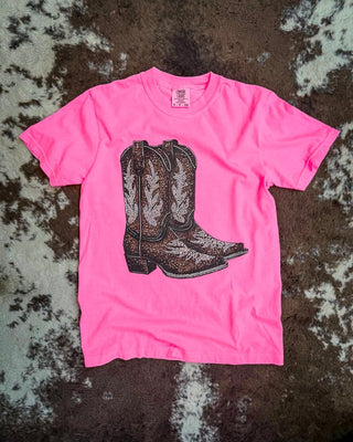A bright pink short-sleeve t-shirt with a graphic of a pair of brown and black patterned cowboy boots, laid flat on a brown and white cowhide rug.