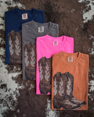 Four folded short-sleeve graphic T-shirts arranged in a fan shape on a brown and white cowhide rug. The shirts are in navy blue, gray, bright pink, and rust orange, each featuring a large western cowboy boot graphic printed on the front.