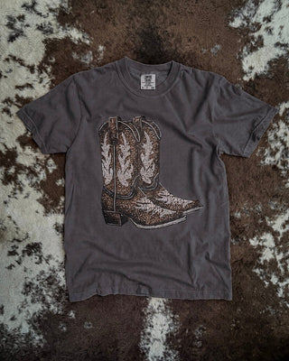 A dark gray t-shirt featuring a graphic print of a pair of western cowboy boots in brown, beige, and white, laid flat on a brown and white cowhide rug.