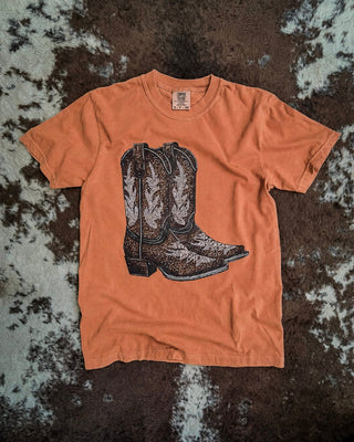 A rust short-sleeve t-shirt with a graphic of a pair of brown and white cowboy boots, laid flat on a brown and white cowhide rug.