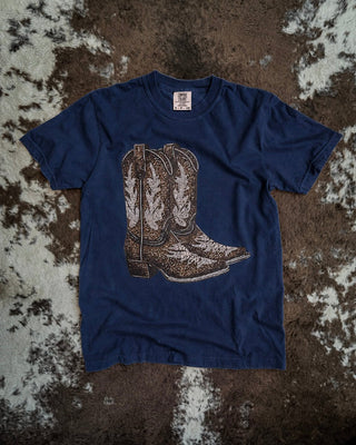 A navy blue short-sleeve t-shirt with a graphic of a pair of brown and white western cowboy boots, laid flat on a brown and white cowhide rug.