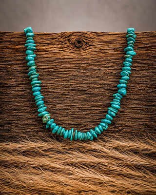 A turquoise beaded necklace made of small, irregular disc-shaped beads, displayed on a rustic brown wooden surface with a textured brown fur-like fabric in the foreground.