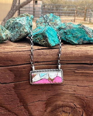 A silver rectangular pendant necklace with a colorful marbled inlay of pink, white, and light blue, resting on a rustic wooden surface with green turquoise rocks in the background.