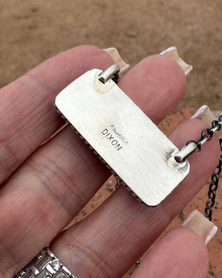 A close-up of a hand holding a silver rectangular pendant necklace engraved with 'Dixon' and 'Sterling'.