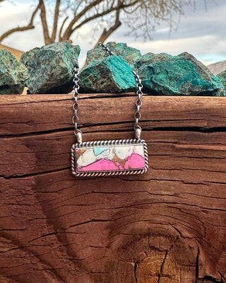 A rectangular pendant necklace with a silver-toned chain and a colorful marbled inlay featuring pink, white, and light blue, displayed on a rustic wooden surface with green rocks in the background.