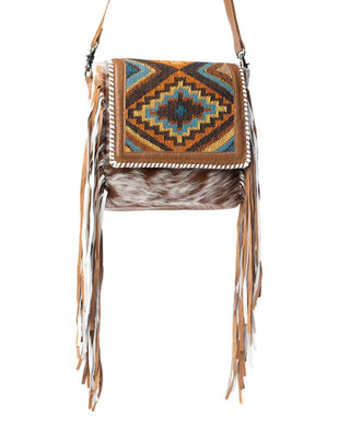 A square crossbody bag featuring a Southwestern geometric patterned flap in brown, orange, and blue, a brown faux fur body, and long tan leather fringe on the sides, shown against a plain white background.