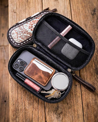 An open black zippered phone wallet organizer case displayed on a rustic wooden table, showing its interior compartments filled with a phone, card holder, keys, lip gloss, and earbuds case. A second closed view of the case with a floral patterned exterior is shown behind it.