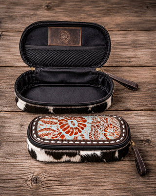 Two zippered pencil cases with cowhide-patterned trim are displayed on a rustic wooden surface. The top case is open, showing a black interior and a brown leather patch with a western-style monogram logo.
