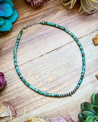A beaded necklace made of turquoise and green beads with a small section of dark metallic beads, displayed on a rustic wooden surface with dried flowers and succulents.