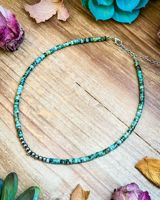 A beaded necklace made of small turquoise-blue and green marbled beads with a silver chain extender, arranged on a rustic wooden surface with dried flowers and greenery around it.