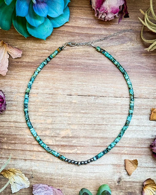 A beaded necklace made of turquoise-blue and green marbled beads with small dark spacer beads, laid flat on a rustic wooden surface with dried flowers and greenery around it.