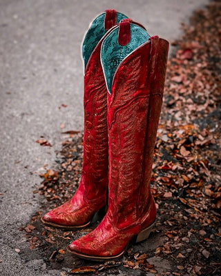 A pair of tall red western cowboy boots with gold-toned embroidery and turquoise insoles, standing on wet pavement surrounded by scattered autumn leaves.