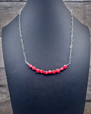 A silver chain necklace with a curved row of glossy red beads, displayed on a black jewelry bust against a dark wood background.