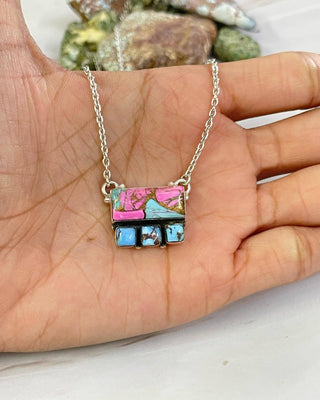 A silver chain necklace with a rectangular pendant featuring colorful mosaic-like segments in pink, blue, black, and green, held in a person's hand.