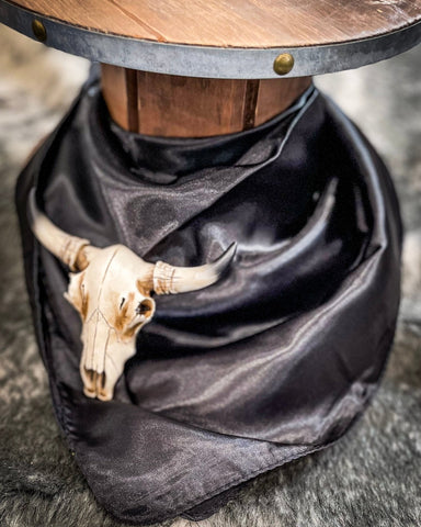 The Wynonna Wild Rag in Black tied around a wooden cable reel with a small cow skull on cowhide.