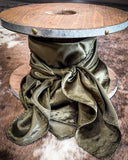 The Wynonna Wild Rag in Olive tied around a wooden cable wheel, resting on a backdrop of cowhide.