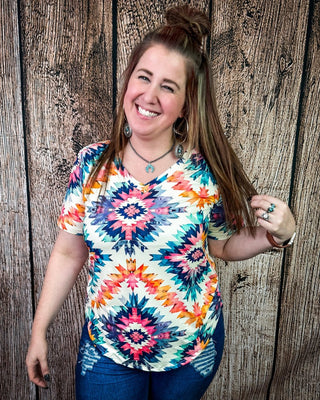 A smiling woman with long brown hair in a high bun wears a colorful short-sleeve V-neck top with a geometric southwestern pattern, paired with blue jeans, standing against a rustic wood plank wall.