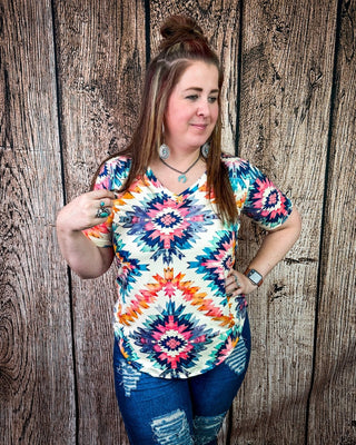 A woman wearing a colorful short-sleeve V-neck top with a vibrant geometric and mandala-like pattern in pink, orange, blue, and white, paired with distressed blue jeans, standing against a rustic wooden plank wall.
