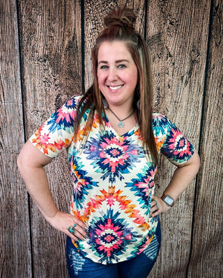 A woman wearing a colorful short-sleeve V-neck top with a vibrant geometric starburst pattern in pink, blue, orange, and yellow on a cream background, paired with blue jeans, posing against a rustic wooden plank wall.