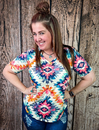 A woman wearing a colorful short-sleeve V-neck top with a geometric tie-dye pattern, paired with blue jeans, posing in front of a rustic wooden plank wall.