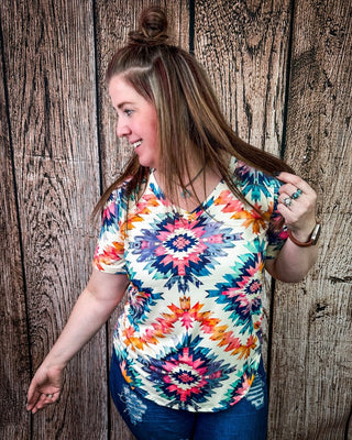 A woman wearing a colorful short-sleeve V-neck top with a vibrant geometric mandala pattern in pink, orange, blue, and yellow on a cream background, paired with blue jeans, standing against a rustic wooden plank wall.