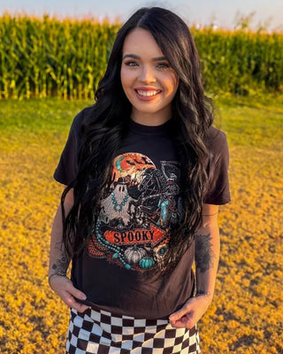 A woman standing in a cornfield wearing a black graphic t-shirt with a spooky skull design and the word 'SPOOKY' on the front, paired with a black and white checkered mini skirt.