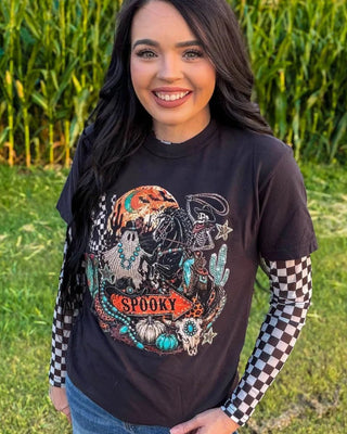 A woman standing outdoors in a grassy field with a cornstalks background, wearing a black graphic t-shirt featuring a spooky western design with a skeleton, cactus, skull, and the word 'SPOOKY', layered over a black-and-white checkered long-sleeve shirt.