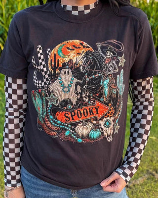 A person wearing a black graphic t-shirt with a spooky Halloween design, layered over a brown and white checkered long-sleeve shirt and blue jeans, standing outdoors on grass.