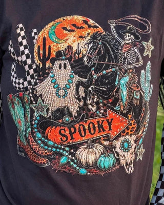 A close-up of a black graphic t-shirt featuring a detailed western-themed 'Spooky' design with a skeleton cowboy, ghost, cactus, skulls, and turquoise accents.