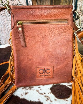 A brown leather crossbody bag with fringe details, a front zip pocket, and a 'Countrywide Co.' logo patch, displayed on a cowhide surface.