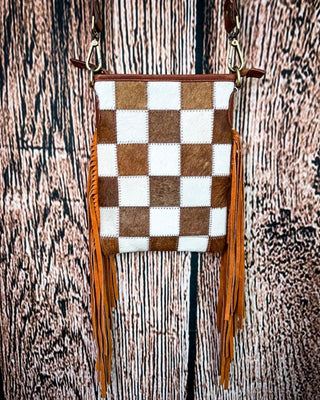 A small crossbody bag with a brown and white checkered hide pattern, brown leather trim, and long orange-brown fringe, hanging against a rustic wood plank background.