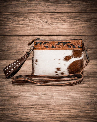 A western-style crossbody clutch bag with a brown and white cowhide body, tooled leather trim, and a matching leather wristlet strap, displayed on a rustic wooden plank background.