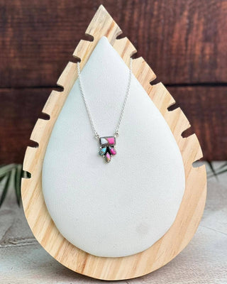 A silver necklace with a small bow pendant featuring pink, green, and blue enamel details, displayed on a white teardrop-shaped stand with a light wood border against a dark wood background.