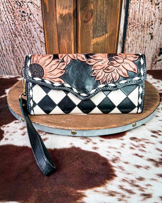 A hand-tooled leather clutch purse with a black and white harlequin diamond pattern, peach floral carvings, and a black wristlet strap, displayed on a rustic wooden stand over a cowhide rug.