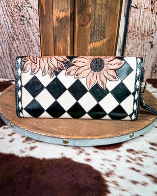 A handcrafted leather clutch wallet featuring a black and white diamond checkerboard pattern, decorated with large tan and black sunflower designs, displayed on a rustic wooden stand.