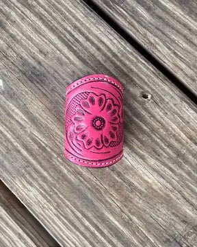 A bright pink leather scarf slide with an embossed floral pattern, resting on weathered gray wooden planks.
