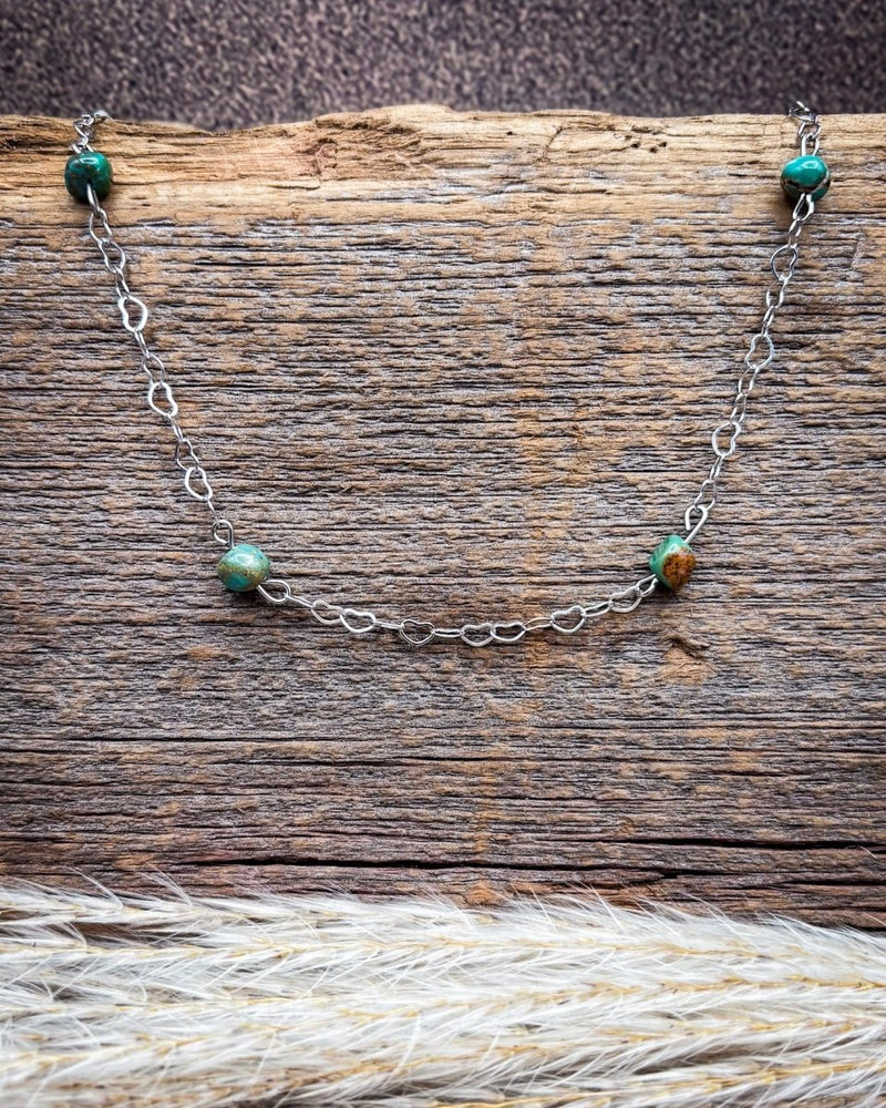 A delicate silver chain necklace with small green and brown beads, displayed on a rustic wooden surface with dried grass in the foreground.