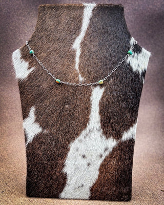 A delicate silver chain necklace with small turquoise and yellow beads, displayed on a brown and white cowhide jewelry bust against a textured brown background.