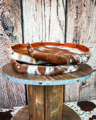 Three stacked cowhide trays with brown and white hair-on-hide tops and brass-tone metal rims, displayed on a rustic wooden pedestal against a wood plank backdrop.