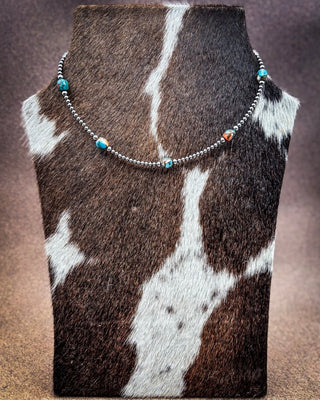 A beaded necklace displayed on a cowhide jewelry bust. The necklace features silver-toned round beads with small turquoise and orange marbled accent beads.