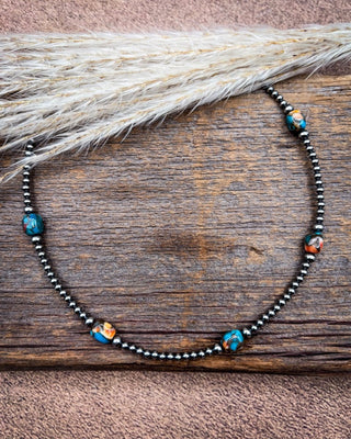 A beaded necklace with dark metallic beads and turquoise, orange, and white marbled accent beads, displayed on a rustic wooden surface with dried pampas grass.