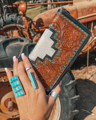 Hand holding a tooled leather wallet with turquoise accents in front of a tractor.