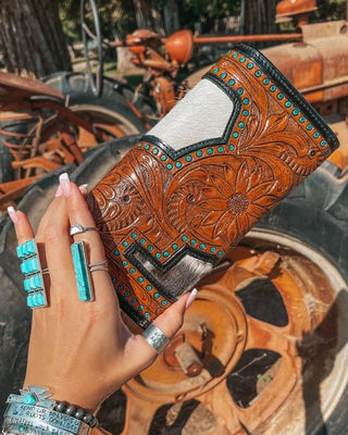 Hand holding a brown leather wallet with intricate designs in front of an old tractor.