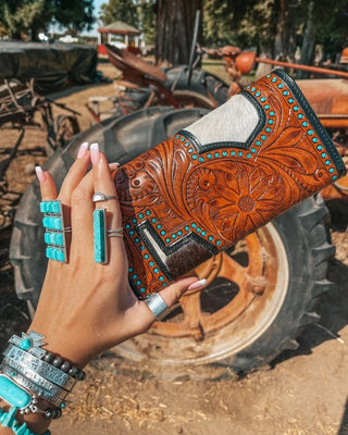 Hand holding a brown leather wallet with turquoise accents in front of a rustic background with a tractor.