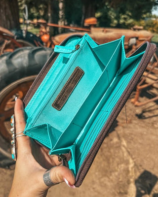 Teal wallet with brown accents held by a hand against a rustic background with a tractor.