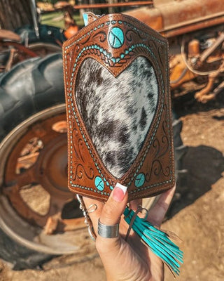 Heart-shaped leather case with cowhide pattern held in front of a rustic background with a tractor.