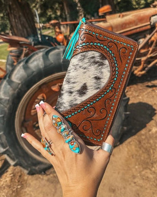 Hand holding a cowhide wallet with intricate designs in front of a rustic background with a tractor.