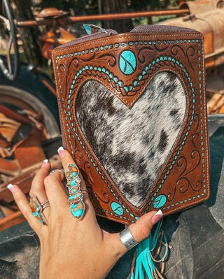 Hand holding a decorative leather wallet with heart design and turquoise accents in front of vintage vehicles.