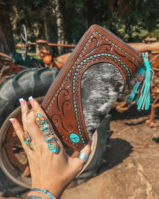 Hand holding a brown leather wallet with turquoise accents in front of a tractor.